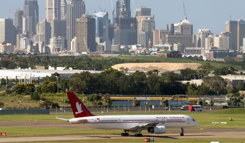 Royal Tongan Airlines plane taxiing on runway with lush greenery and the Sydney city skyline in the background.